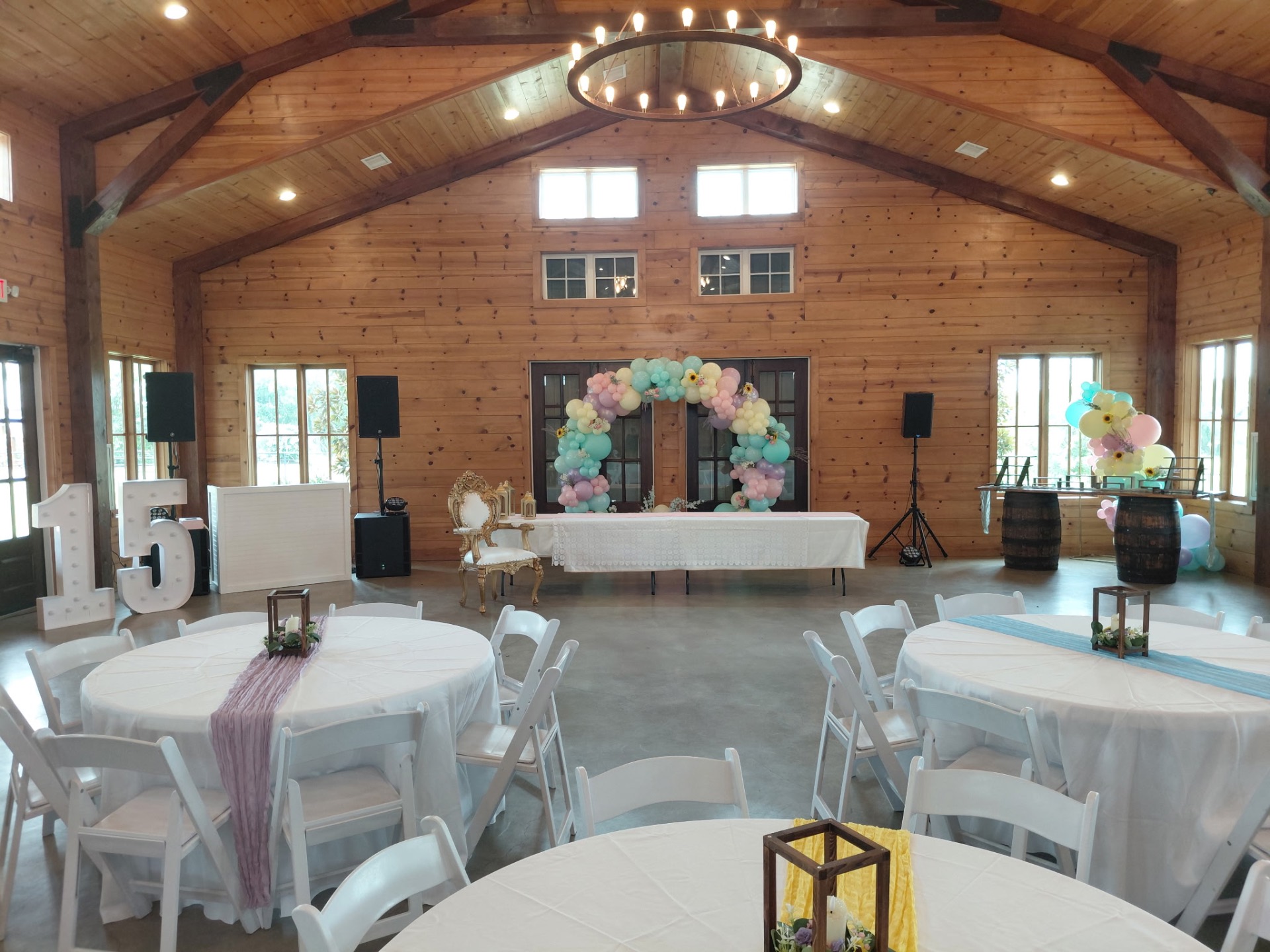 Quinceañera venue with pastel balloon arch, marquee 15 letters, gold throne, and round guest tables