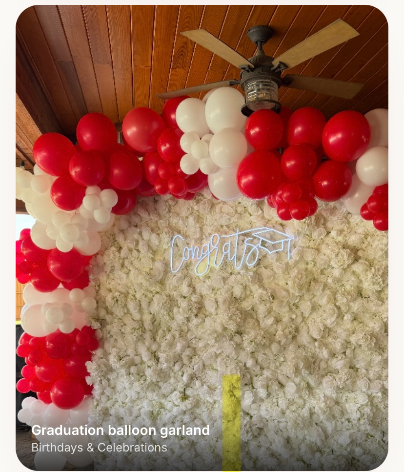 Red and white organic balloon garland framing a white floral wall with a Congrats neon sign